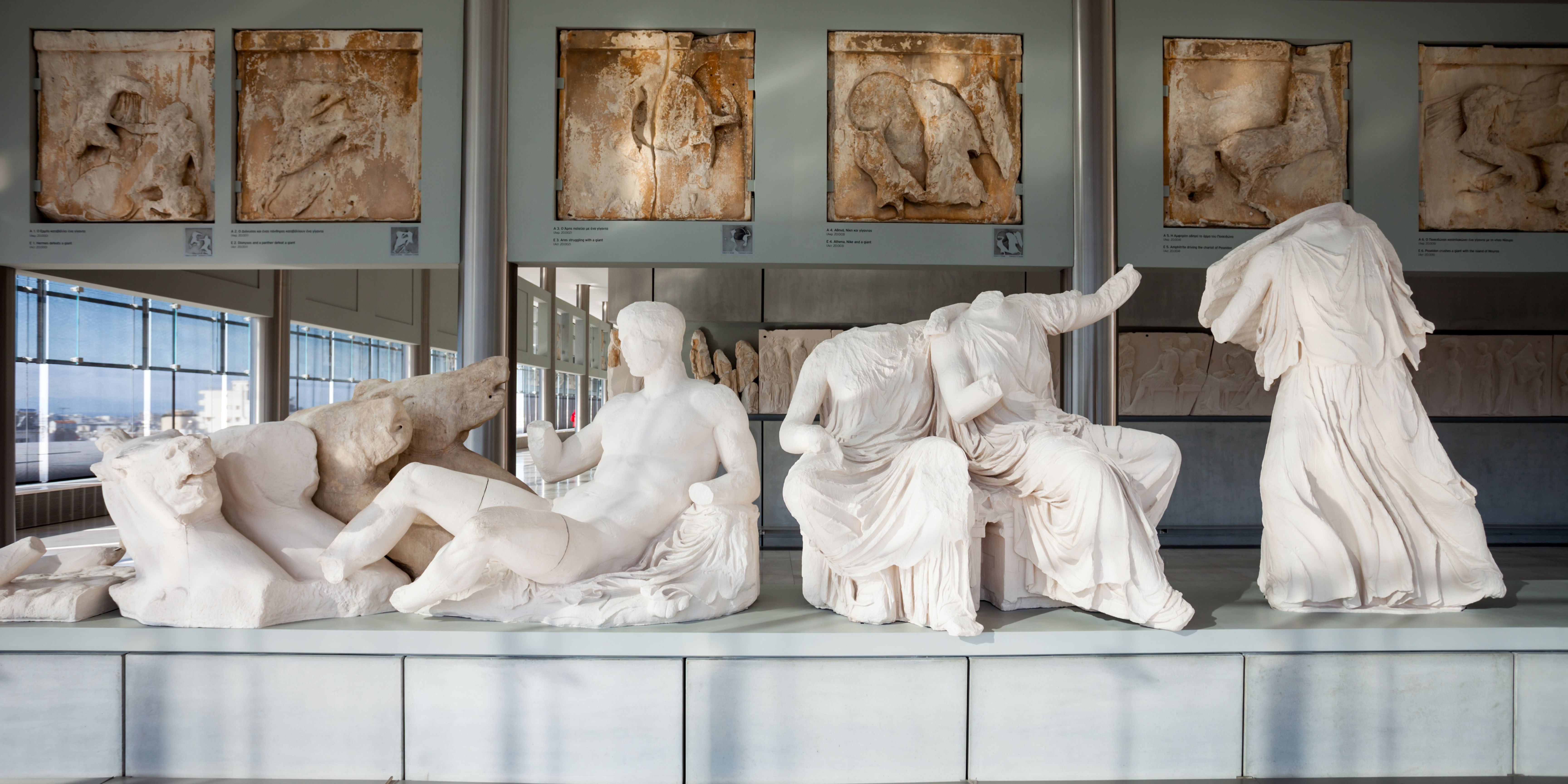 Sculptures and friezes on display in a museum with large windows in the background.